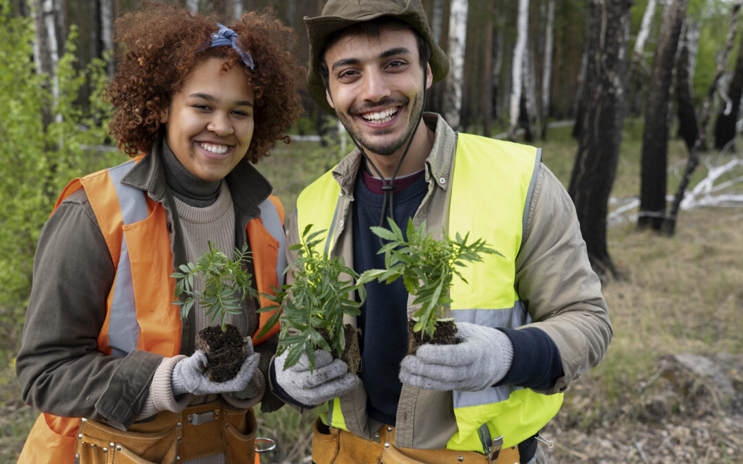 Deux jeunes adultes reconvertis aux métiers de l'écologie, et équipés pour le jardinage, sourient en tenant dans leurs mains des plantes prêtes à être plantées.